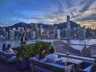 Sunset over Victoria Harbour, with the Hong Kong Island skyline seen from a rooftop terrace, guests seated with glasses in hand