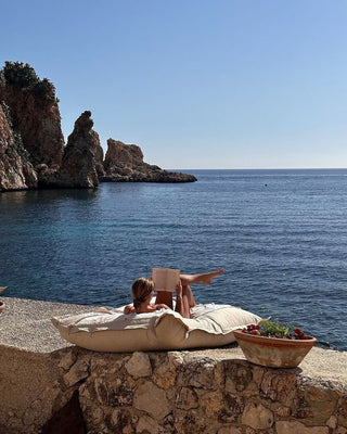 A peaceful coastal retreat, with a woman lounging on a cushioned seat by the sea, reading a book under the warm Mediterranean sun. Rugged cliffs and the sparkling blue water create a perfect backdrop for relaxation and escape